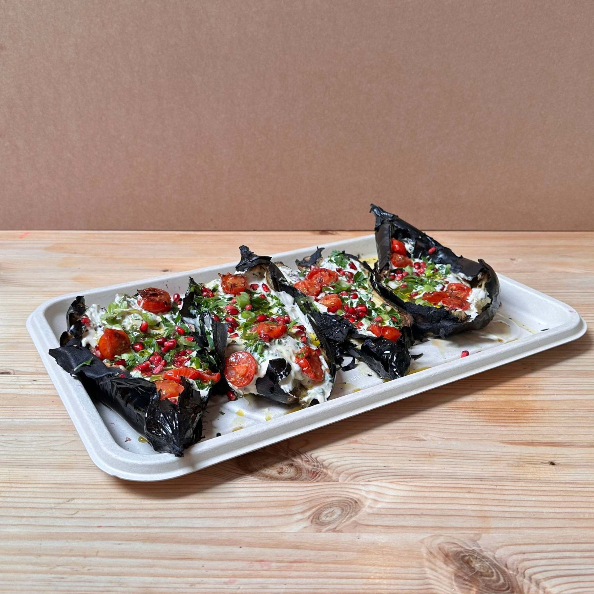 A platter of whole chargrilled smoked aubergines covered with tahini and tomato salsa, grilled cherry tomatoes, zhug, pomegranate seeds, scallions and parsley
